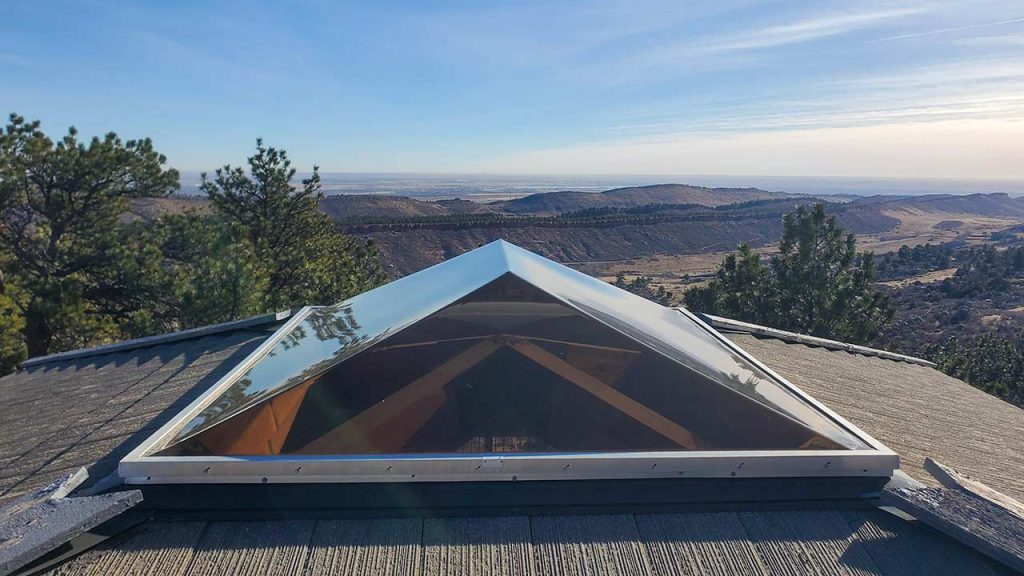 A bronze acrylic skylight on a roof overlooks a vast landscape. Trees surround the scene, with rolling hills under a clear blue sky in the background.