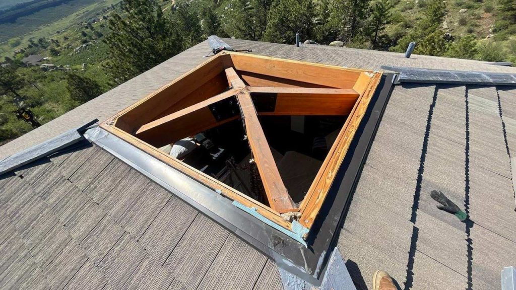 View of a roof with a large skylight opening exposing wooden beams. Green trees and hills can be seen in the background. A sense of open space.