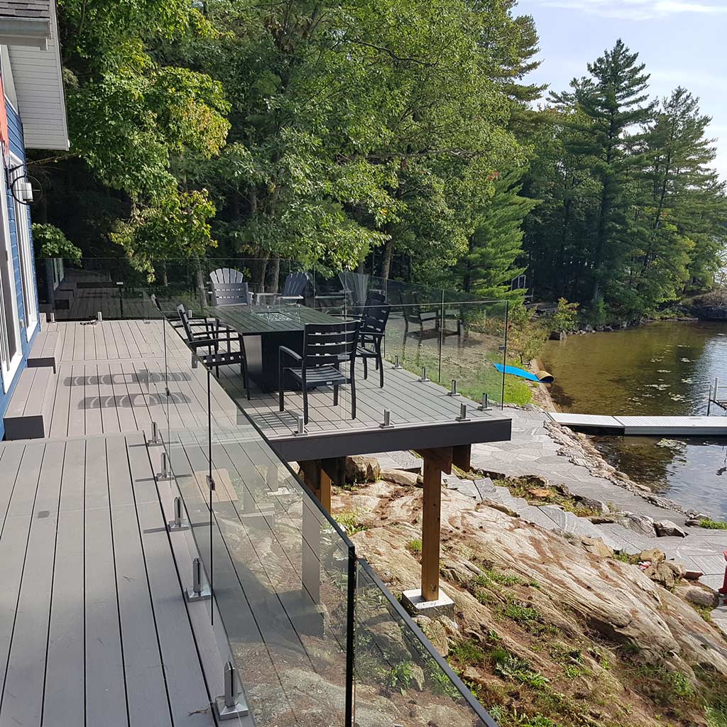 Modern lakefront deck with glass railings, black dining set under trees, overlooking calm water and stone pathway. Serene and inviting outdoor space.