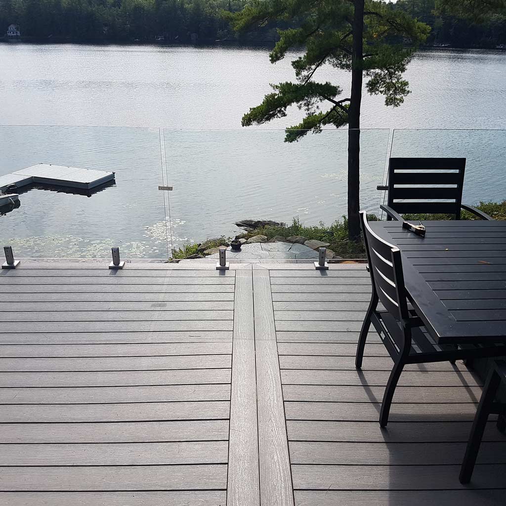 A serene lakeside view from a wooden deck featuring a dark patio table with chairs, glass railing, and a floating dock. Calm and peaceful atmosphere.
