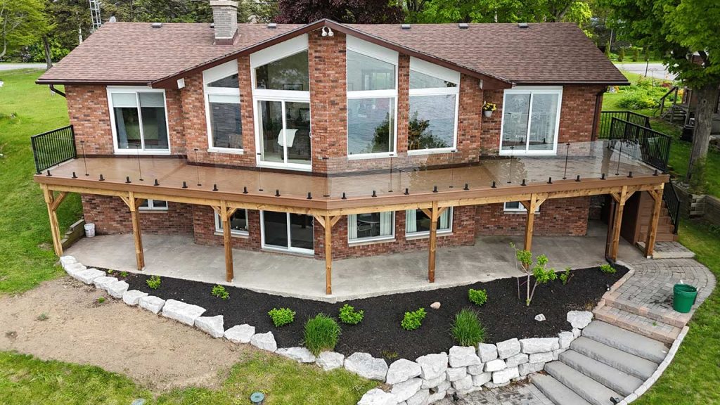 Two-story brick house with large windows and a wraparound wooden deck with a glass railing , surrounded by neatly landscaped garden with white stones and green plants.