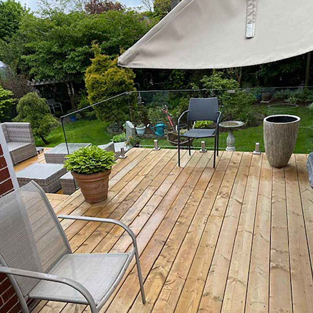 Wooden patio deck with a glass railing and metal chairs, large potted plant, and a patio umbrella partially covering greenery. Relaxing and inviting outdoor setting.
