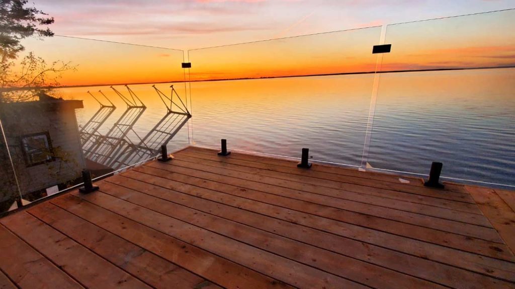Sunset view of a serene lake from a glass-railed wooden deck. The sky is a gradient of orange and pink, reflecting warmly on the calm water.