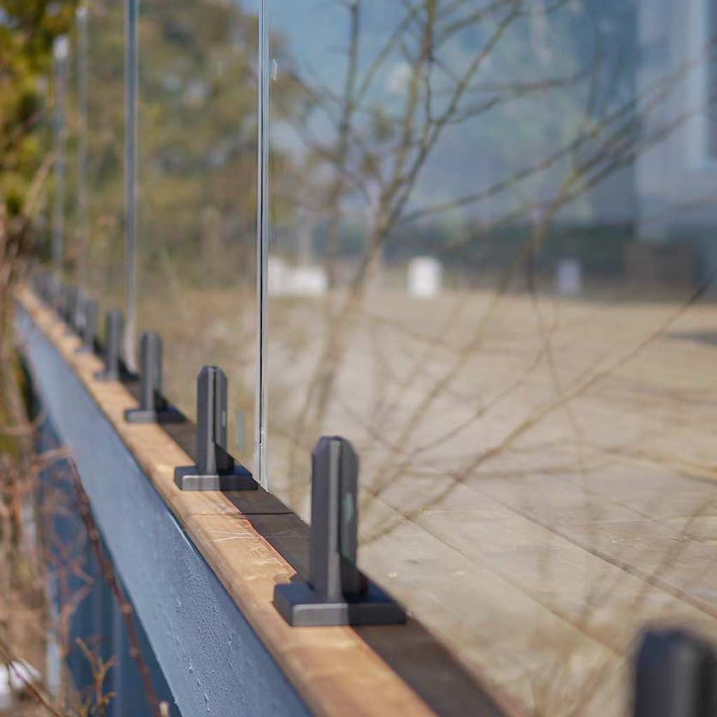 Glass railing with black bases line a wooden edge, overlooking a blurred background of trees and a building, creating a serene, modern feel.