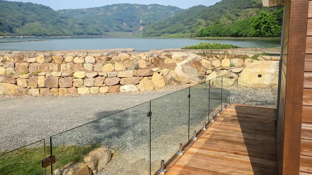 A serene lakeside view with lush green hills in the background. A wooden deck with glass railing overlooks a stone wall and gravel path, evoking tranquility.