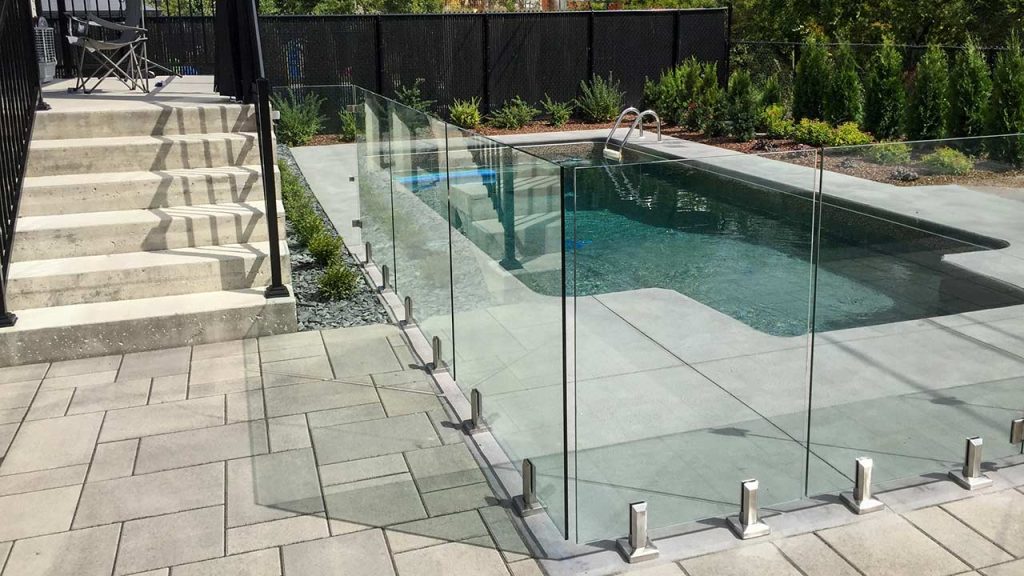 A modern backyard with a rectangular swimming pool surrounded by a clear glass railing. Concrete steps and patio lead up to the pool area, with greenery in the background.