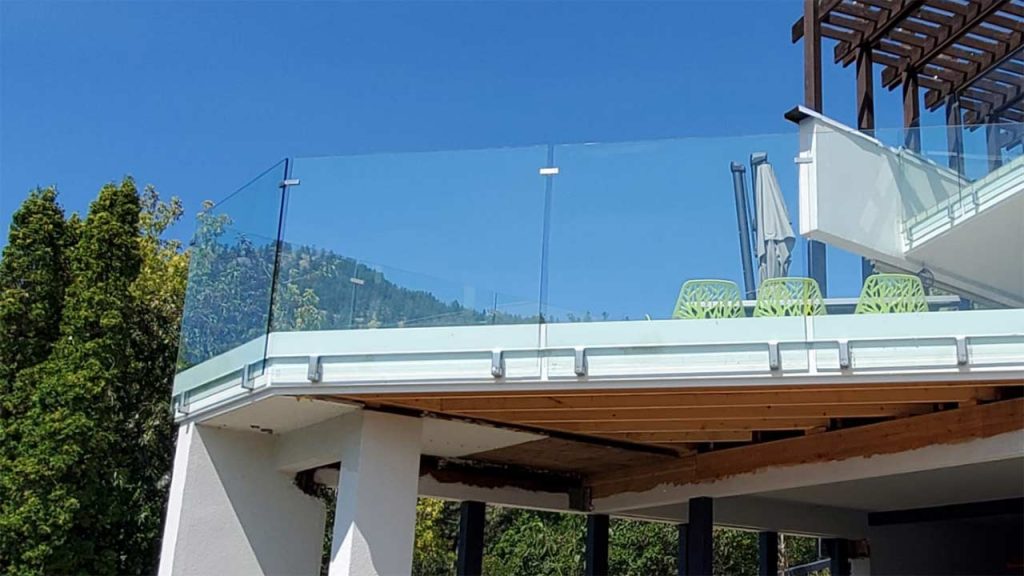 Modern balcony with glass railing, overlooking lush green trees and hills under a bright blue sky. Green chairs and umbrella create a serene, open atmosphere.