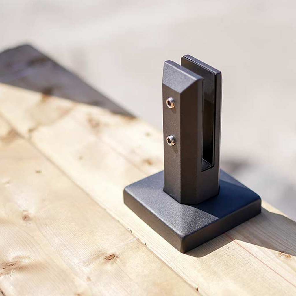 A metal fence spigot is mounted on a sunlit wooden deck. The spigot has a sleek, angular design, casting a shadow. The scene feels calm and contemporary.