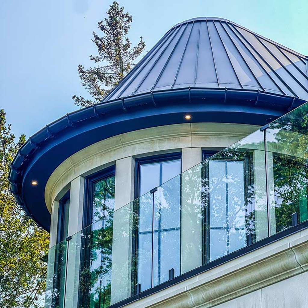 A modern building with a circular glass railing around a balcony and a cone-shaped metal roof is surrounded by tall trees, evoking an elegant and serene atmosphere.