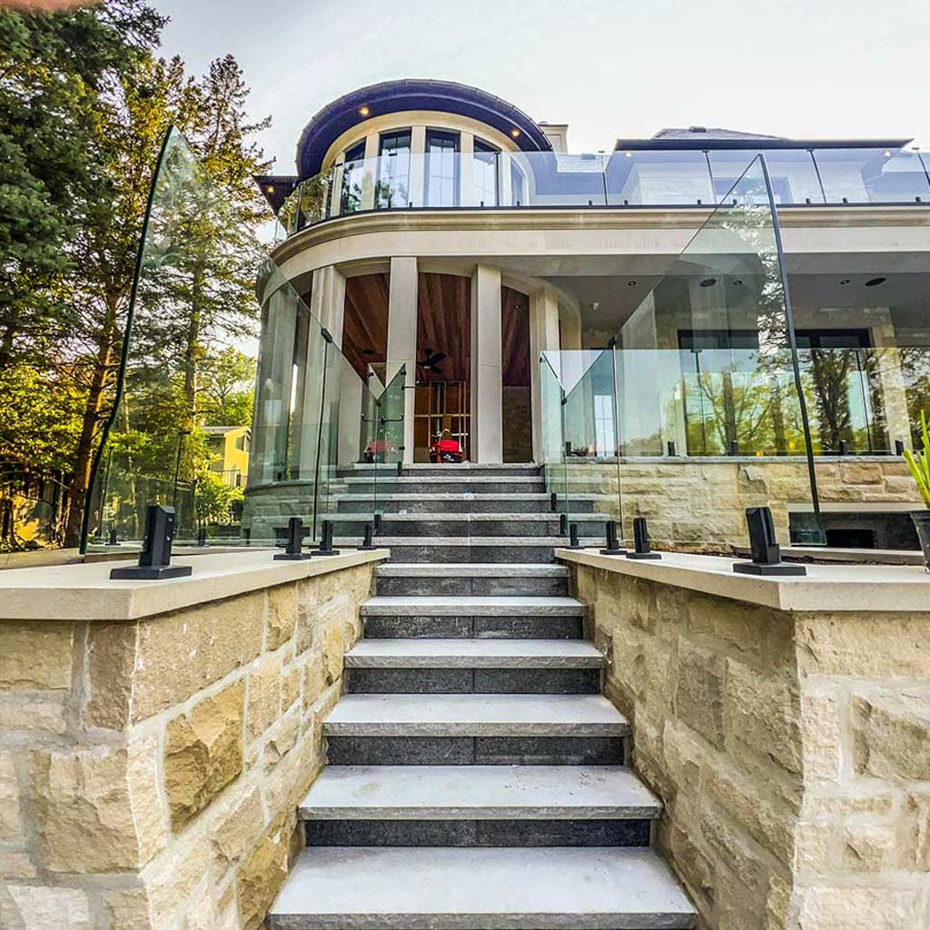 Modern two-story house with large glass windows and stone facade. Stone steps lead up to a glass railing entrance, surrounded by trees and greenery.
