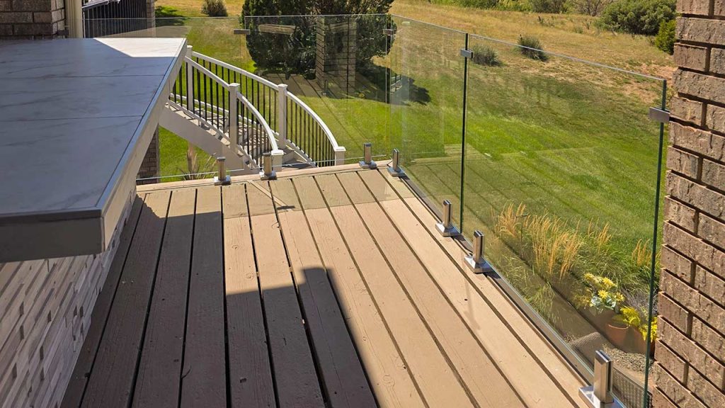 Sunset over a serene beach, viewed through a glass railing. The sky is a gradient of orange to blue, with calm waters reflecting the sun's glow.