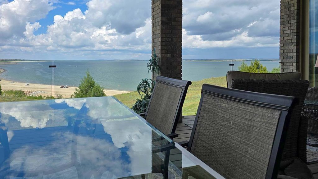 A glass table on a balcony with a glass railing reflects a cloudy sky. The setting overlooks a tranquil beach and ocean, creating a serene and peaceful atmosphere.
