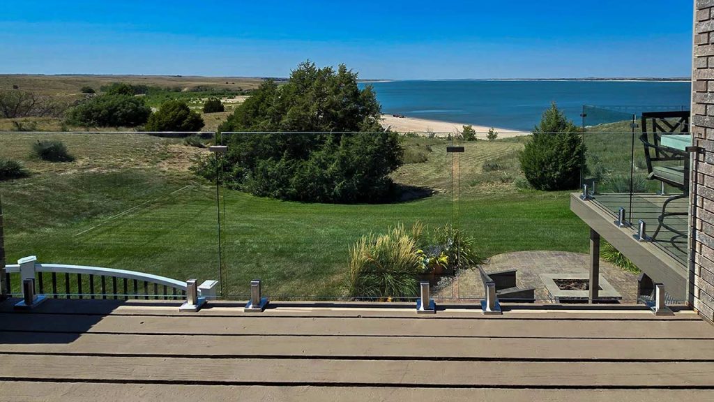 A scenic view from a balcony with glass railing shows a grassy yard leading to a beach and expansive blue lake, bordered by trees under a clear sky.