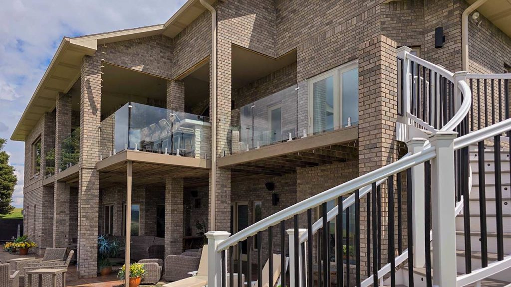 A two-story brick house with a balcony with a glass railing and patio. White and black railings line the staircase. Modern design, inviting outdoor seating.