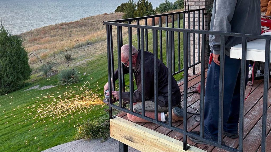 Man kneeling on wooden deck is removing a metal balcony railing with a grinder as sparks fly and by a lakeside view. Another person stands nearby. Scene conveys focus and craftsmanship.