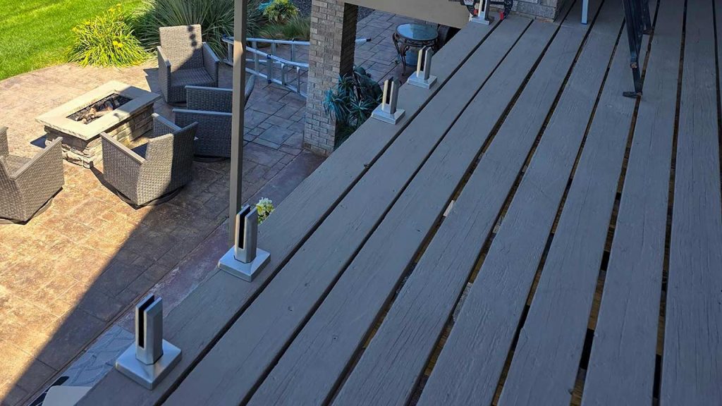 Elevated balcony with wooden decking, and glass railing spigots overlooking a stone-paved area. Modern wicker chairs encircle a fire pit. Bright, sunny ambiance.
