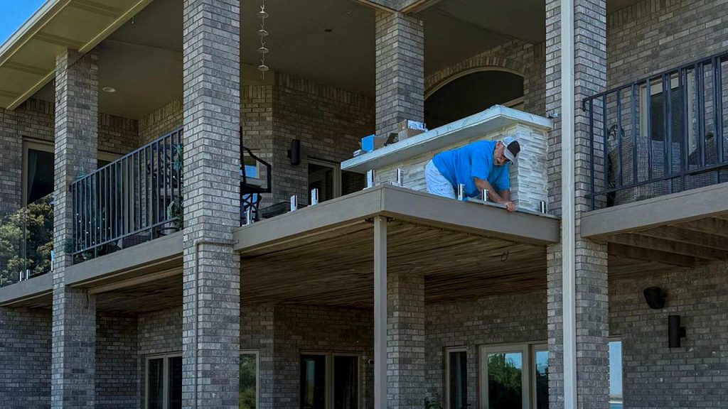 A man in a blue shirt installs a glass railing on a brick house’s balcony. The building has multiple stories with balconies, conveying a work-in-progress tone.