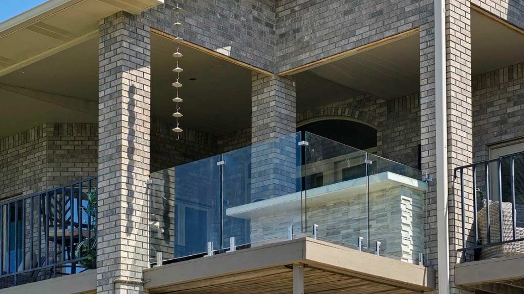 Modern house exterior with a corner balcony featuring glass railings and brick walls. A wind chime hangs, creating a serene and elegant atmosphere.