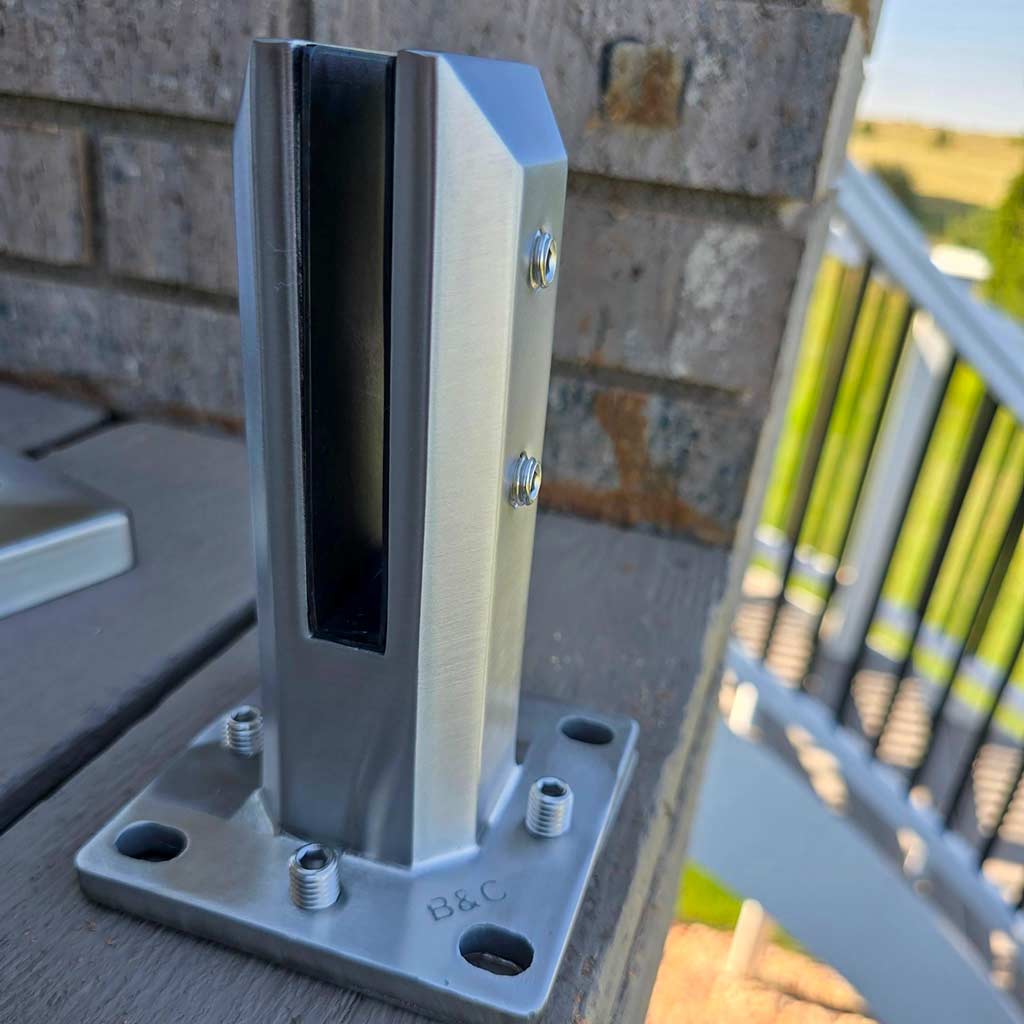 Close-up of a shiny stainless steel glass railing post mounted on a wooden deck. The background shows a blurred outdoor scene with greenery.