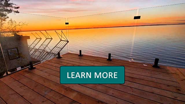unset view of a serene lake from a glass-railed wooden deck. The sky is a gradient of orange and pink, reflecting warmly on the calm water. Learn More overlay.