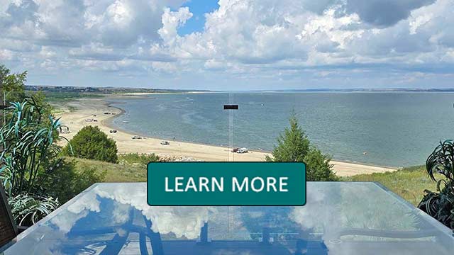 A scenic view through a flass railing of a sandy beach and calm lake under a partly cloudy sky. Greenery borders the beach, with a glass table reflecting the clouds. Tranquil atmosphere.Learn More overlay.