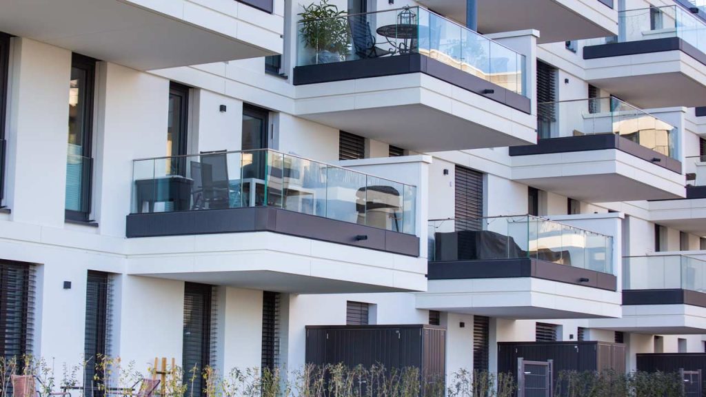 Modern apartment building with white facade and multiple balconies with glass railings. Some balconies have plants and outdoor furniture, conveying a clean, urban atmosphere.