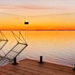 Wooden deck overlooking a calm lake at sunset, with glass railings and three stacked chairs. The sky is a blend of orange and purple hues, creating a serene atmosphere.
