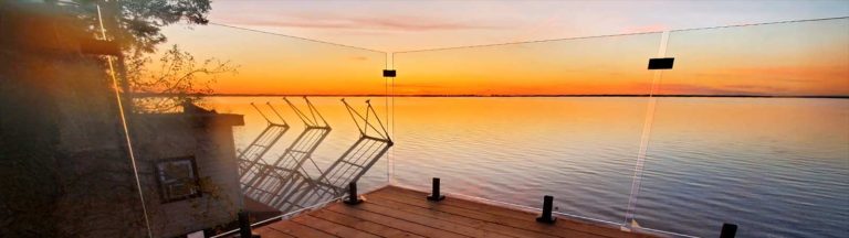 Wooden deck overlooking a calm lake at sunset, with glass railings and three stacked chairs. The sky is a blend of orange and purple hues, creating a serene atmosphere.