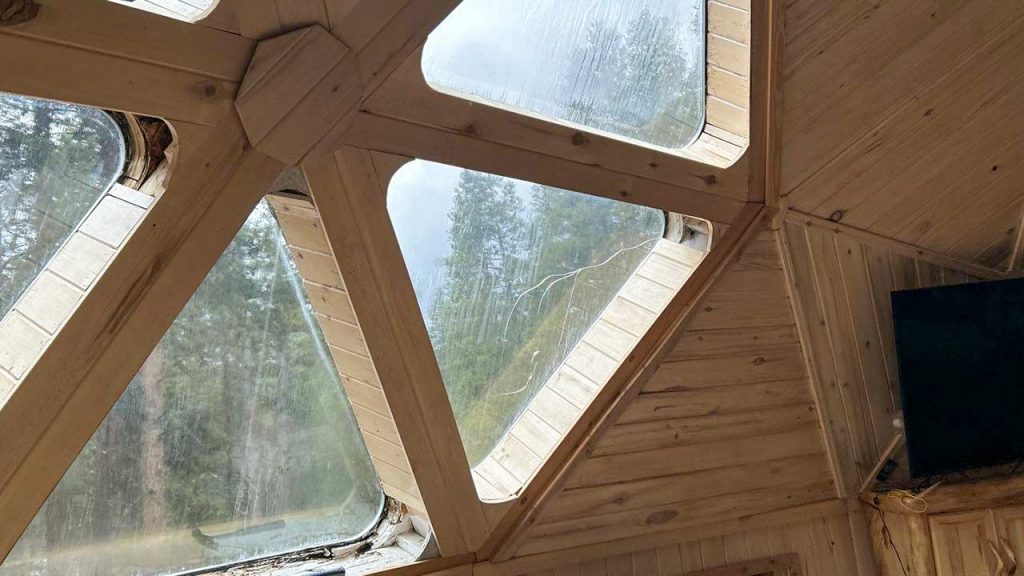 Geometric wooden ceiling with triangular windows, showing a cloudy sky and green trees outside. The cozy interior exudes a warm, rustic feel.