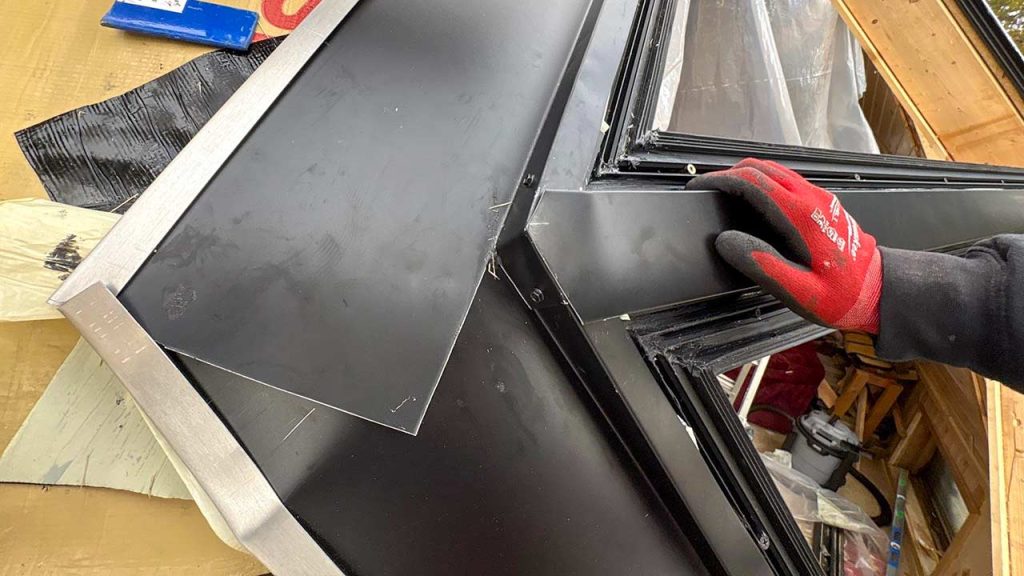 A person wearing red gloves installs black metal flashing on a roof corner, with wooden beams visible in the background, suggesting construction work.