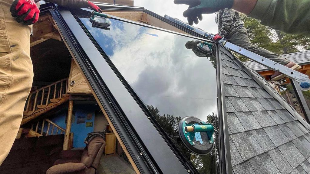 Workers install a reflective, triangular glass panel on a cabin roof, surrounded by trees. The panel reflects the cloudy sky. Mood is industrious and focused.