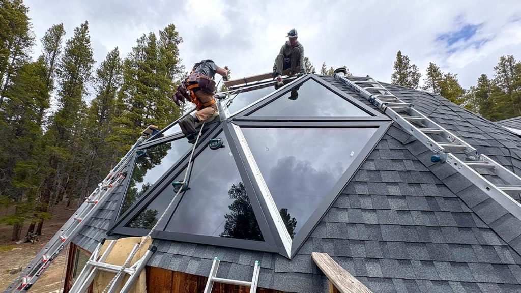 Workers install large triangular glass panels on a geodesic dome roof amidst a forest setting, using ladders. The scene conveys teamwork and innovation.
