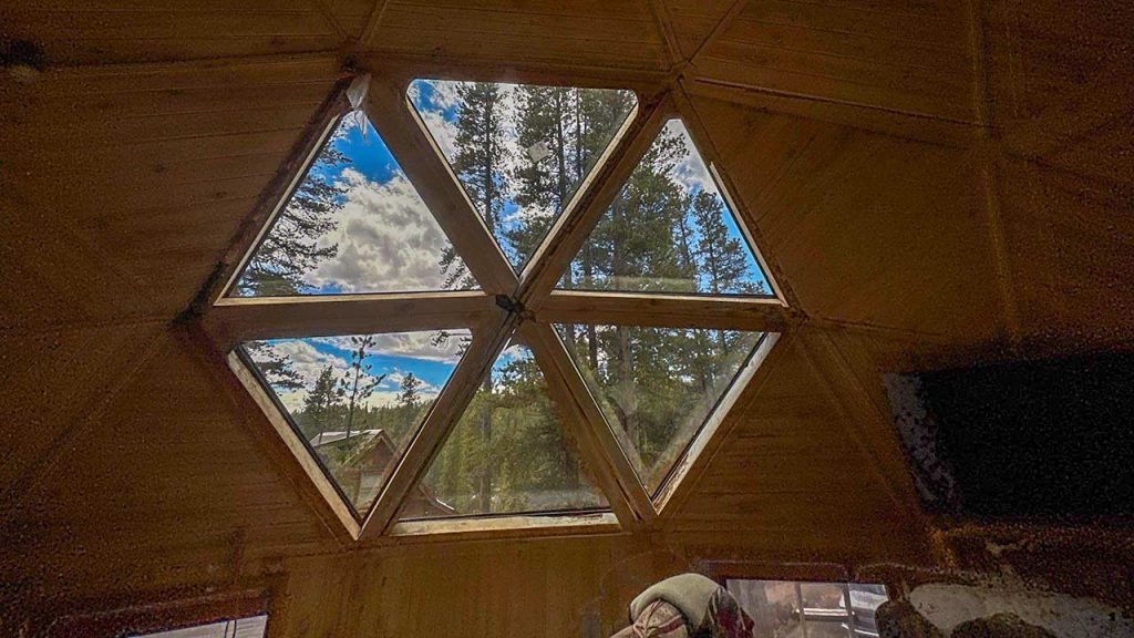 Geometric wooden ceiling with large triangular windows showing a view of blue sky and tall pine trees, creating a serene and natural atmosphere.