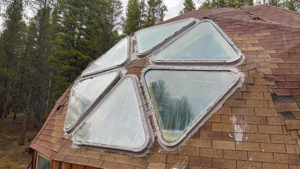 Geodesic dome roof with triangular windows, surrounded by dense forest. The structure's shingles contrast with the overcast sky, evoking a rustic feel.