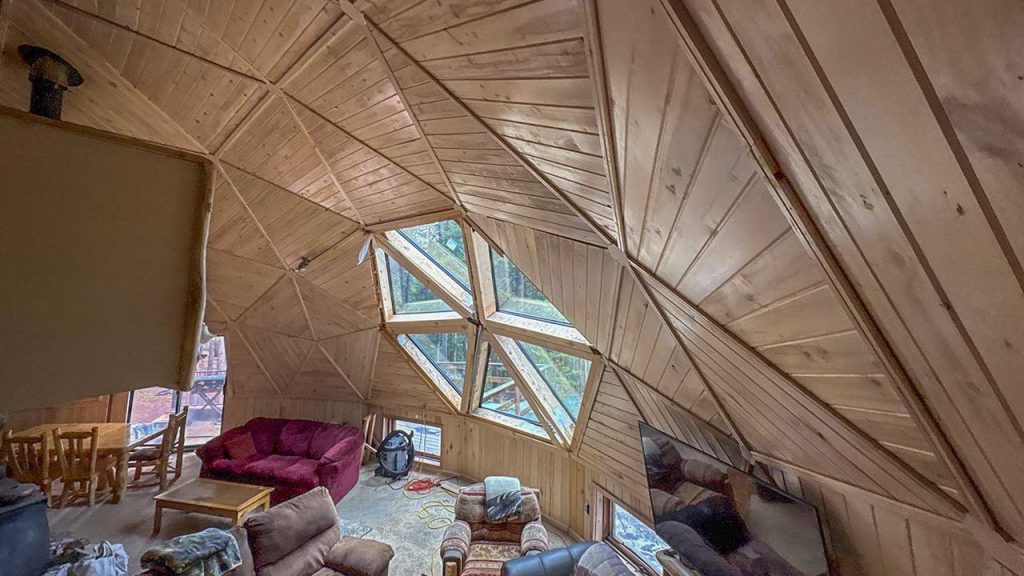 Geodesic dome interior with wooden paneling, showcasing hexagonal skylight. Cozy seating area with sofas and a table, exuding warmth and comfort.
