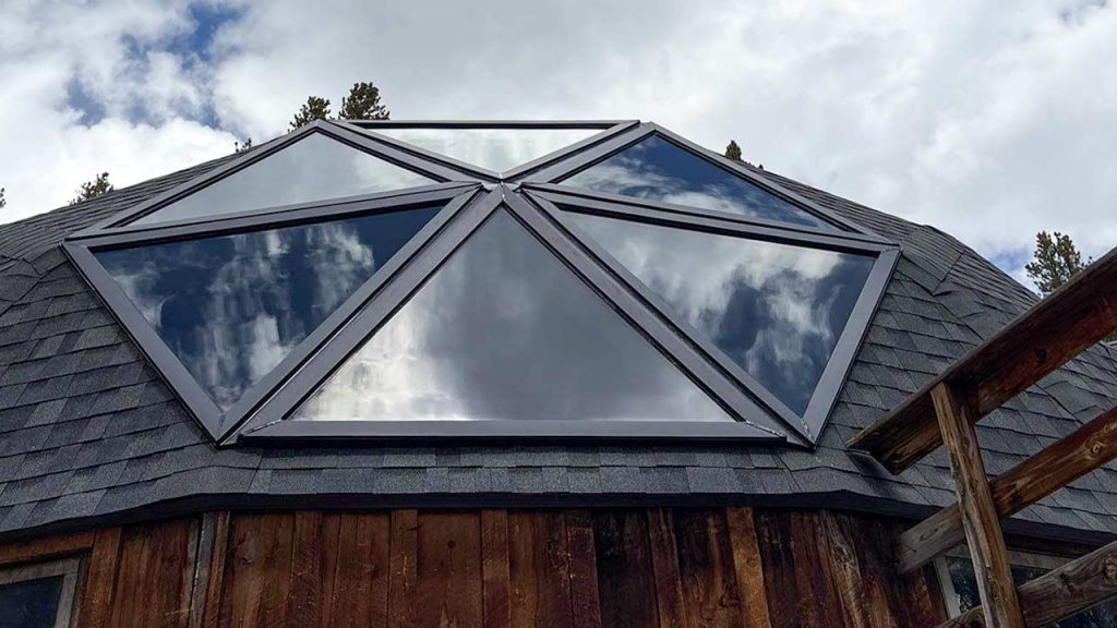 Geodesic dome skylight with triangular glass panels reflects a cloudy sky. Rustic wooden walls and shingled roof create a natural, peaceful atmosphere.