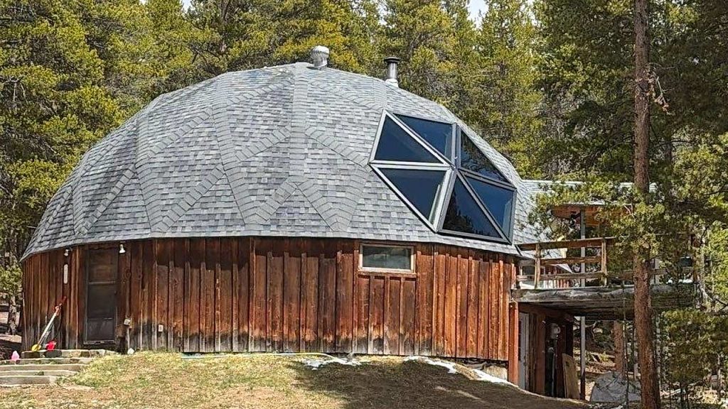 A geodesic dome house with a wooden exterior blends into a forest setting. Large hexagonal skylight on the roof reflect sunlight. The scene feels tranquil and natural.