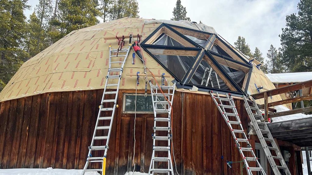 A geodesic dome cabin amidst snow-covered trees with ladders leaning against its wooden exterior. People work on installing large triangular windows.