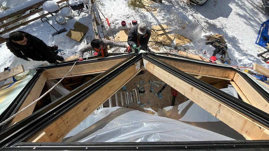 Three people work on constructing a Mags Bar hexagonal skylight on roof in a snowy area. The scene conveys teamwork and effort on a clear winter day.