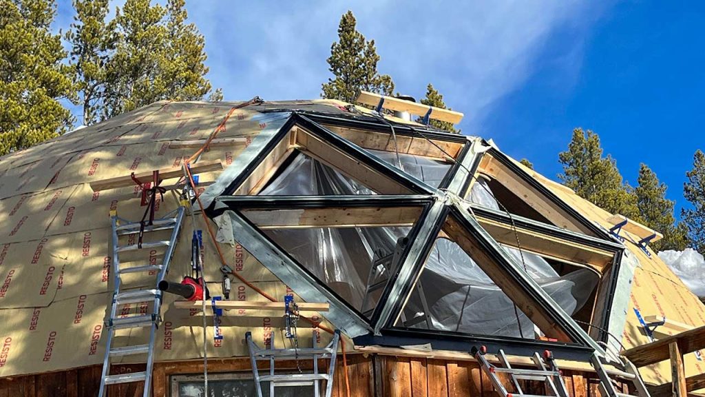 A geodesic dome home under construction with a large, hexagonal Mags Bar skylight frame. Surrounded by pine trees and blue sky, it conveys a sense of openness and innovation.