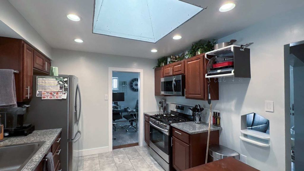 Modern kitchen with dark wood cabinets, stainless steel appliances, and tiled floor. Recessed lighting and a skylight illuminates the space, giving it a bright, airy feel. An open doorway reveals an office area with a chair and computer.